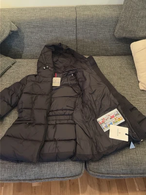 Svart dunjacka från Moncler med huva Dam - Snygg svart dunjacka från Moncler med huva och midjeresår. Jackan har quiltad design, dragkedja framtill och två sidofickor. Klassisk Moncler-logga på ärmen och innerficka. Perfekt för kalla vinterdagar och riktigt varm.