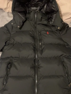 Svart dunjacka från Polo Ralph Lauren - Snygg svart dunjacka från Polo Ralph Lauren med huva och dragkedja framtill. Jackan har två sidofickor med dragkedja och en bröstficka. Klassisk röd logga broderad på bröstet. Perfekt för kalla vinterdagar och riktigt skön att bära.