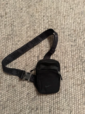 Svart liten axelväska från Nike - Säljer en svart liten axelväska från Nike med justerbart band där det står NIKE i svart text. Väskan har dragkedja upptill, ett extra fack framtill och är tillverkad i slitstark nylon. Perfekt för att bära det viktigaste och har en sportig look.