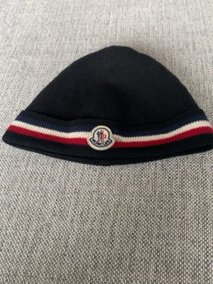 Tricolor moncler mössa  - Snygg svart stickad mössa från Moncler med klassisk randig kant i blått, vitt och rött. Mössan har Moncler-loggan framtill och är tillverkad i mjukt material som håller dig varm. Perfekt accessoar för kyliga dagar eller bara för stilens skull ,hör av er vid frågor 🤞