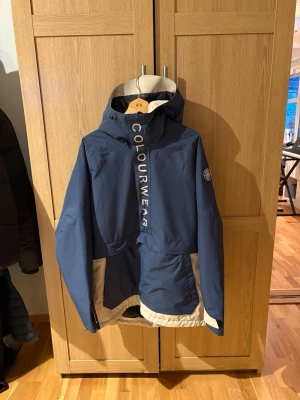 Blå skidjacka från CLWR - Snygg blå vindjacka från ColourWear med beige detaljer och stor huva. Jackan har dragkedja framtill, stora fickor och logga på ärmen. Materialet är vindtätt och lätt, perfekt för blåsiga dagar. CLWR-tryck på huva och bröst ger en sportig look. Passform baggie 