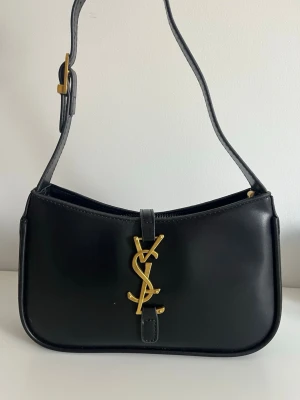 Svart läder axelväska med logga i guld - Så snygg handväska med guldig logga. Det finns en innerficka med dragkedja samt ett extra förlängnings band om man önskar att förlänga remmen. Väskan stängs med en dragkedja och klasp. 😊 