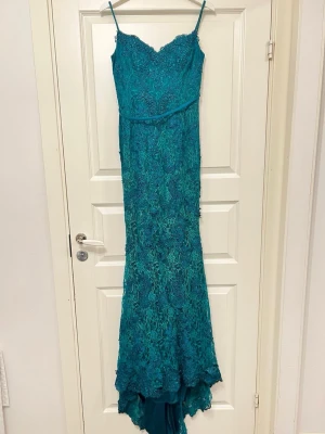 Turkos långklänning med spetsdetaljer - Säljer en elegant turkos långklänning med smala axelband och v-ringad urringning. Klänningen har vackra spetsdetaljer över hela plagget och en lätt utsvängd nederdel. Perfekt för bal eller festliga tillfällen.