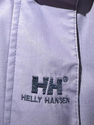 Jacka från Helly Hansen - Snygg lilablå skaljacka från Helly Hansen med mörkblå ärmar och detaljer. Jackan har huva, dragkedja framtill och broderad logga på bröstet. Materialet är slitstarkt och vattenavvisande, perfekt för blåsiga dagar. Justerbar nederkant och muddar för extra komfort.