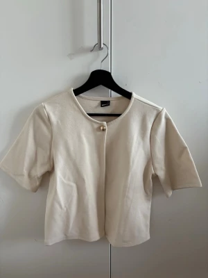 Beige kortärmad bolerotopp med knapp - Jättesöt kofta från gina med knapp. Använd endast en gång och nästan i nyskick. Passar bra över ett linne eller liknande.