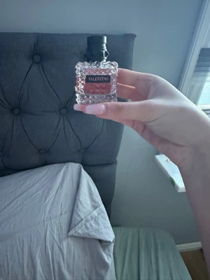 Valentino parfym med kristallflaska - Lyxig parfym från Valentino i en fyrkantig, transparent kristallflaska med nitar och rosa doftvätska. Flaskan har en svart kork och elegant etikett framtill. Perfekt för dig som vill ha en exklusiv och trendig doft på hyllan.