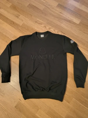 Svart Moncler sweatshirt - Svart sweatshirt från Moncler med broderad logga och text på bröstet samt patch på ena ärmen. Klassisk rund halsringning och ribbade muddar vid ärmslut och nederkant. Perfekt för en clean och stilren look.