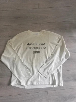 Vit långärmad topp Acne Studios - Storlek S. Vit långärmad topp från Acne Studios med tryck 'Acne Studios STOCKHOLM 1996' på bröstet. Klassisk rund halsringning och relaxed fit. Perfekt till jeans eller kjol för en clean och stilren look.