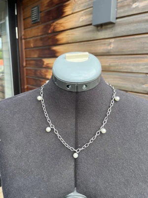 Silverfärgat halsband med pärlor - Kedjehalsband i silverfärg med små vita pärlor jämnt fördelade längs länkarna. Halsbandet har en enkel och stilren design som passar till många olika looks. Perfekt för dig som gillar minimalistiska smycken med en touch av elegans.