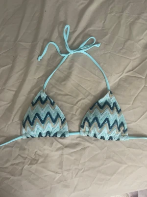 Blå zigzag bikiniöverdel med knyt - Snygg trekantsbikini med zigzag-mönster i olika blå toner och vitt. Överdelen har tunna band som knyts i nacken och ryggen för justerbar passform. Perfekt för strandhäng eller pooldagar.