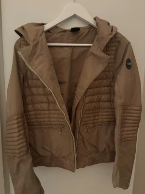 Beige pufferjacka från colmar - Snygg beige pufferjacka från colmat med kort, figurnära passform och quiltade paneler. Jackan har dragkedja framtill, två sidofickor och colmat-logga på ärmen. Perfekt för kalla dagar och ger en trendig look till din outfit.