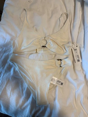 Beige bikini med ringdetaljer - Trendig beige bikini från H&M med ribbad struktur och snygga ringdetaljer både på toppen och trosan. Både överdel och underdel har en twist framtill och smala axelband. Perfekt för sommarens strandhäng!