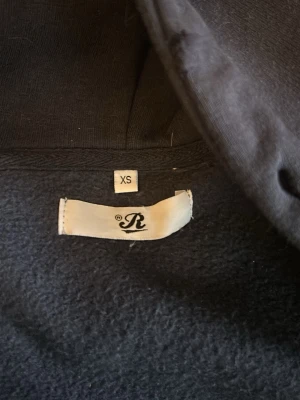 Svart hoodie från Russedress XS - Mörkblå hoodie med dragkedja från Russedress i storlek XS. Hoodien har huva, två fickor framtill och ribbade muddar vid ärmslut och nederkant. Broderad Russedress-logga på ena ärmen. Perfekt för chill dagar och streetwear-stil.