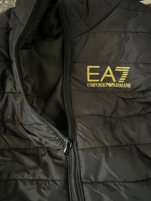 Svart dunväst från Emporio Armani EA7 - Snygg svart dunväst från Emporio Armani EA7 med gul logga på bröstet. Västen har quiltad design, dragkedja framtill och hög krage. Perfekt för lager-på-lager och ger en clean streetwear-look. Materialet är lätt och smidigt, vilket gör den enkel att bära.