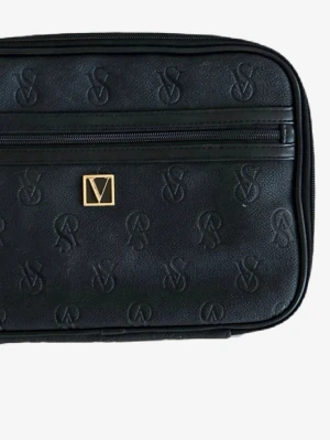 Svart necessär från Victoria Secret - Stilren svart necessär från Valentino med präglad logotyp över hela väskan och guldfärgad metallogo framtill. Väskan har en rektangulär form och ett ytterfack med dragkedja. Tillverkad i konstläder med slät finish.
