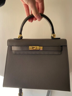 Grå Kelly väska 👜 - Snygg grå H Kelly inspirerad handväska i strukturerat läder med guldfärgade metalldetaljer och klassiskt vridlås. Väskan har ett topphandtag och en stilren, elegant form som gör den till en riktig ikon. Perfekt för dig som vill ha en exklusiv accessoar med lyxig känsla.