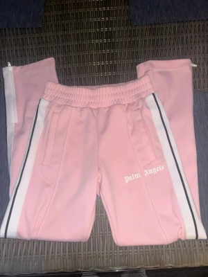 Rosa mjukisbyxor från Palm Angels - Snygga rosa mjukisbyxor från Palm Angels med vita och svarta ränder längs sidorna. Byxorna har resår i midjan, sidofickor och dragkedja vid benslut. Perfekta för en chill och trendig look.