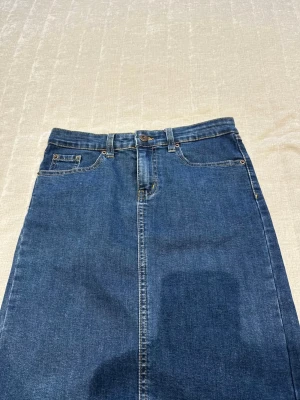 Klassisk blå jeanskjol - Snygg blå jeanskjol med femficksmodell, bälteshällor och knappstängning framtill. Kjolen har en rak passform och slits bak för extra rörelsefrihet. Perfekt att styla med en t-shirt eller hoodie för en avslappnad look.