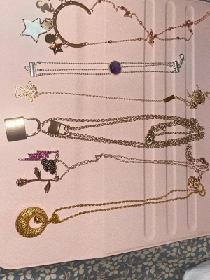 Mixade halsband med hängen i guld och silver - Säljer ett set med flera halsband i guld- och silverfärg. Halsbanden har olika hängen som stjärna, måne, lås, blomma, cirkel och en lila sten. Några hängen har detaljer som små stjärnor och blixtar. Perfekt för dig som gillar att variera din stil med unika accessoarer.