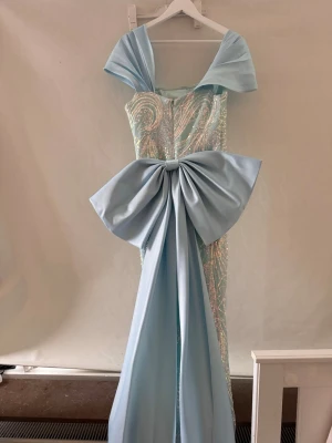 Ljusblå långklänning med stor rosett - Elegant ljusblå långklänning med offshoulder-design och stor dramatisk rosett i midjan. Klänningen har glittriga detaljer och ett figurnära snitt som ger en lyxig känsla. Perfekt för bal eller festliga tillfällen. Använd 1 gång. Storlek 38, säljs för 800 kr.