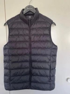 Marinblå dunväst från Uniqlo - Snygg marinblå dunväst från Uniqlo med quiltad design och hög krage. Västen har dragkedja framtill och är lätt vadderad för extra värme. Perfekt att slänga över en hoodie eller tröja när det är kyligt ute.