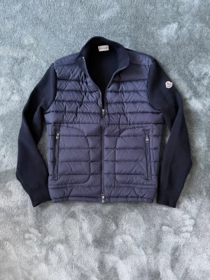 Moncler cardigan - Snygg mörkblå dunväst från Moncler med stickade ärmar och quiltad framsida. Västen har två dragkedjefickor och en hög krage. Klassisk Moncler-logga på ena ärmen och stilren design som passar perfekt till lager-på-lager.