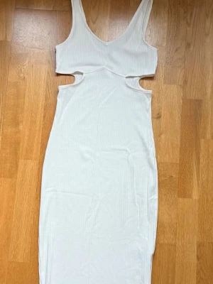 Vit ribbad lång klänning med cut-outs - Trendig vit ribbad klänning med cut-outs i midjan och v-ringad hals. Klänningen är ärmlös och har en kroppsnära passform som framhäver siluetten. Perfekt för sommarens alla tillfällen.