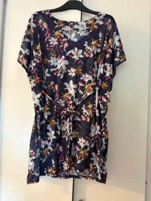 Marinblå blommig strandklänning - Snygg marinblå strandklänning med blommigt mönster i vitt, rosa och gult. Klänningen har korta ärmar, rund halsringning och ett knytband i midjan för en avslappnad passform. Perfekt för sommardagar på stranden.