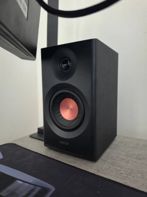 Edifier svart trådlös högtalare - Snygg svart trådlös högtalare från Edifier med modern design. Högtalaren har en rund kopparfärgad detalj i mitten av basen och en diskant ovanför. Tillverkad i robust plast och metall, perfekt för gaming eller musik vid datorn.