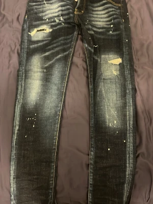 Mörkblå slitna jeans med färgstänk dsq - Säljer ett par mörkblå jeans med slitningar och trendiga färgstänk. Jeansen har skinny fit, tydliga slitningar på låren och knäna samt kontrastsömmar. Perfekta för dig som gillar en edgy streetstil.
