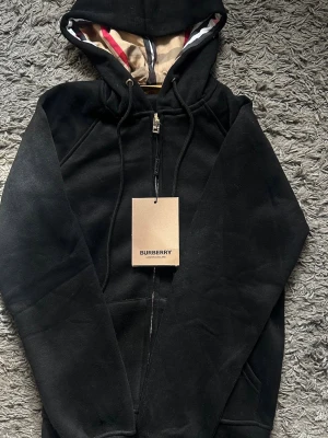 Burberry zip - Snygg svart hoodie från Burberry med dragkedja och klassisk huva. Insidan av huvan har Burberrys ikoniska rutmönster och detaljer i beige. Hoodien har fickor på sidorna och guldfärgade metalldetaljer på snörena. Perfekt för en stilren och avslappnad look. Storlek S