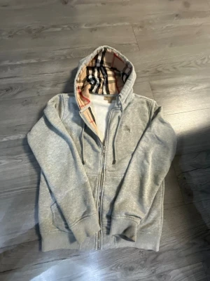 Grå zip hoodie från Burberry - Snygg grå zip hoodie från Burberry med klassiskt rutigt foder i huvan. Hoodien har dragkedja framtill, två fickor och ribbade muddar vid ärmar och nederkant. Perfekt för dig som gillar stilrena plagg med en twist. Materialet känns mjukt och bekvämt.