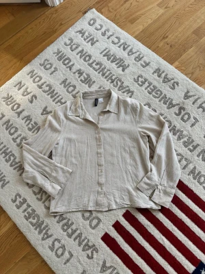 Beige linneskjorta från H&M - Stilren beige skjorta i linne från H&M med klassisk krage och knäppning framtill. Skjortan har långa ärmar med manschett och knappdetalj. Perfekt för en avslappnad men snygg look.