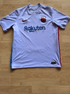 Ljus lila FC Barcelona matchtröja Nike - Snygg  FC Barcelona matchtröja från Nike med klubbmärke och Rakuten-tryck på bröstet. Tröjan har blå och röda detaljer på sidorna samt UNICEF-tryck på ryggen. Tillverkad i lätt och ventilerande material, perfekt för fotboll eller träning. Storlek 158-170 (XL)