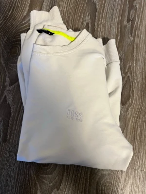 Vit hoodie från BOSS - Snygg vit hoodie från BOSS med diskret broderad logga på bröstet. Klassisk modell med ribbade muddar vid ärmslut och nederkant. Tillverkad i mjukt material som känns skönt mot huden. Perfekt för en clean och stilren look.
