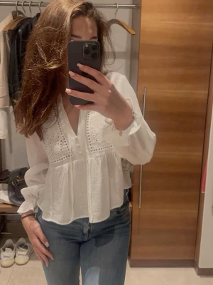 Vit blus med broderade detaljer - Snygg vit blus med v-ringning och broderade detaljer upptill. Blusen har långa ärmar med resår vid ärmslut och en luftig, lätt struktur. Perfekt till jeans eller kjol för en fräsch look.
