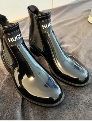 Hugo skor - Snygga svarta Chelsea boots från Hugo i glansigt lackmaterial. Klassisk siluett med resår på sidorna och tydlig logga framtill. Perfekta för dig som vill sticka ut med en modern och trendig look.