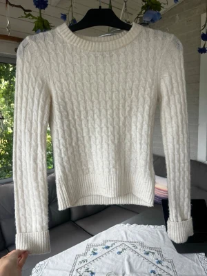 Vit kabelstickad tröja - Kabelstickad vit tröja med rund halsringning och långa ärmar. Tröjan har ribbade muddar vid ärmslut och nederkant, samt en klassisk och tidlös design som passar till mycket. Materialet ser mjukt och mysigt ut, perfekt för kyligare dagar eller en sen sommarkväll