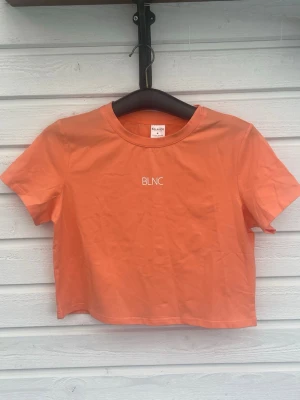 Orange croppad t-shirt från BLNC - Trendig orange croppad t-shirt från BLNC med enkel vit logga på bröstet. T-shirten har korta ärmar och rund halsringning. Perfekt för dig som gillar en avslappnad och modern stil. Aldrig använd eller tvättad