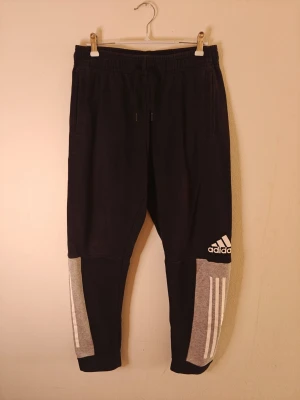 Svarta Adidas mjukisbyxor med grå detaljer - Säljer ett par svarta mjukisbyxor från Adidas med grå paneler och vita ränder på benen. Byxorna har resår i midjan med dragsko, två sidofickor och Adidas-logga på vänster ben. Perfekta för chill eller träning.