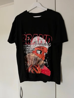 Svart t-shirt med grafiskt tryck - Cool svart t-shirt med stort grafiskt tryck i rött, vitt och beige på både fram- och baksida. Trycket visar en tecknad figur med pengar och texten 'BRAND Presents'. T-shirten har normal passform och är tillverkad i mjuk bomull.