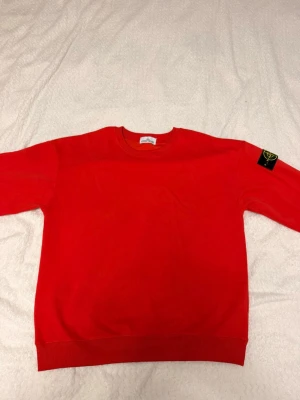 Röd sweatshirt från Stone Island - Snygg röd sweatshirt från Stone Island med klassisk rund halsringning och ribbade muddar. Tröjan har den ikoniska Stone Island-loggan på vänster ärm och är tillverkad i mjukt bomullsmaterial. Perfekt för en avslappnad och trendig stil.