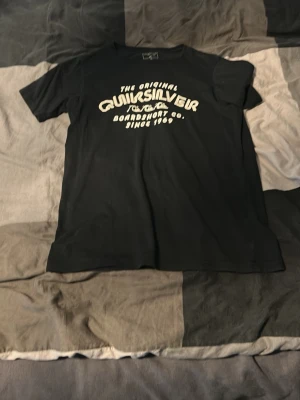 Svart Quiksilver t-shirt med tryck - Snygg svart t-shirt från Quiksilver med vitt tryck på bröstet där det står 'The Original Quiksilver Boardshort Co. Since 1969'. Klassisk passform och rund halsringning. Perfekt för en avslappnad streetwear-look.