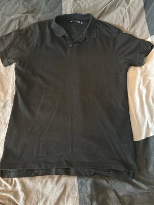 Svart pikétröja med krage - Klassisk svart pikétröja med krage och knappar framtill. Tröjan har korta ärmar och är tillverkad i mjukt bomullsmaterial. Enkel och stilren design som passar till det mesta.