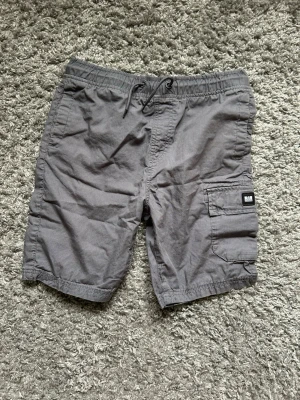 Grå cargoshorts från Weekend Offender - Säljer ett par grå cargoshorts från Weekend Offender med snörning i midjan och praktiska sidofickor. Shortsen har en klassisk cargomodell med en patch på benet och är tillverkade i bomull. Perfekta för en avslappnad stil.