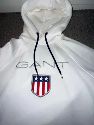 Vit hoodie från GANT med emblem - Snygg vit hoodie från GANT med broderad logga och ett stort emblem med stjärnor och ränder på bröstet. Klassisk huva med svarta dragsnören och mjukt material som känns riktigt skönt. Perfekt för dig som gillar stilrena och sportiga plagg. Tröjan är aldrig använd och storlek XS.