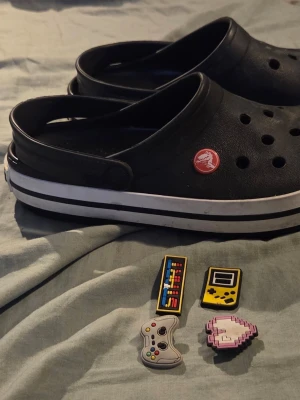 Svarta Crocs med Jibbitz-charms - Säljer ett par svarta Crocs med vit rand runt sulan och flera färgglada Jibbitz-charms med gaming- och matmotiv. Tofflorna har klassisk perforerad ovandel och justerbar hälrem. Perfekta för dig som vill ha bekväma och personliga skor.