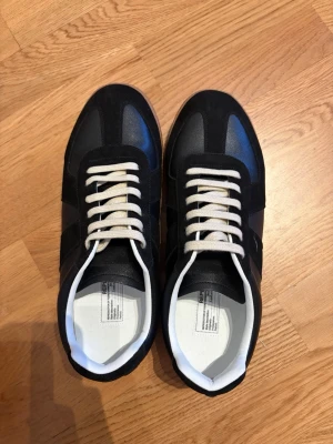 Svarta Maison Margiela skor - Stilrena svarta sneakers med vita skosnören och vit insida. Ovandelen är i en mix av mocka och slätt läder, med klassisk gummisula i brunt. Snygg retroinspirerad design som passar till det mesta. Perfekta för dig som gillar minimalistisk och tidlös stil.