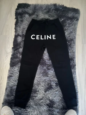 Svarta mjukisbyxor från Celine - Snygga svarta mjukisbyxor från Celine med vit logga framtill. Byxorna har resår i midjan med dragsko, två sidofickor och ribbade muddar vid bensluten. Perfekta för en chill och trendig look.