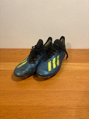 Adidas blå fotbollsskor med gula detaljer - Säljer ett par Adidas fotbollsskor i blått med gula ränder och detaljer. Skorna har snörning, låg profil och mönstrad ovandel för bättre bollkontroll. Yttersulan har dobbar för bra grepp på planen. Perfekta för dig som vill sticka ut på matchen!